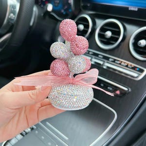 Könnte beinhalten: Ein Auto-Lufterfrischer mit einem funkelnden, mit Edelsteinen besetzten Design. Die Basis hat eine abgerundete Form, und die Oberseite weist eine Ansammlung von rosa und silbernen Kugeln auf. Ein rosa Band ist um die Basis gebunden.