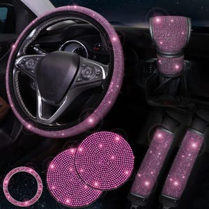 Könnte beinhalten: Ein rosa Auto-Innenraum-Zubehör-Set. Das Set enthält eine Lenkradabdeckung, einen Schalthebelbezug, Sicherheitsgurtbezüge, zwei Getränkehalter und einen Startknopfring, alle mit funkelnden Strasssteinen verziert.