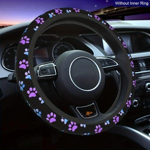 May include: Black steering wheel cover with a pattern of purple and blue paw prints. The cover is designed to fit over a standard steering wheel. The image shows the cover installed in a car, with the dashboard and other interior features visible.