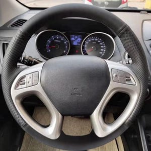 Può includere: Un volante in pelle nera con dettagli argentati e comandi integrati. Il volante ha un'impugnatura traforata e circonda un mozzo centrale con la scritta "AIRBAG". Gli indicatori del cruscotto sono visibili sullo sfondo.