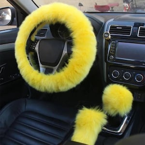 May include: A bright yellow, fluffy steering wheel cover and matching accessories in a car interior. The set includes a steering wheel cover, gear shift cover, and a center console knob cover. The car's interior is black.