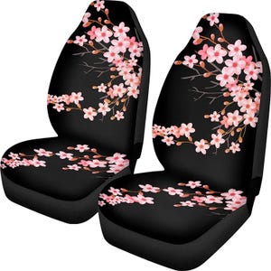 May include: Two black car seat covers with a pink cherry blossom design. The floral pattern features delicate pink flowers and brown branches, creating a contrast against the black background. The seat covers are designed to fit standard car seats.