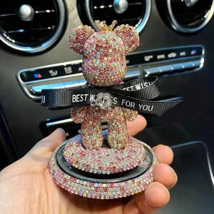 May include: A bedazzled teddy bear car accessory, covered in pink, red, and clear rhinestones. The bear wears a gold crown and has a black ribbon with the text "BEST WISHES FOR YOU". The accessory sits on a circular base.