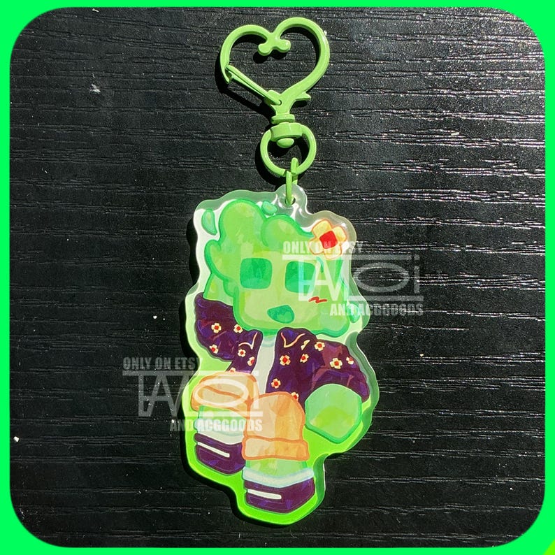 K&ouml;nnte beinhalten: Ein gr&uuml;ner Acryl-Schl&uuml;sselanh&auml;nger mit einer Cartoon-Figur mit gr&uuml;nem Teint, die eine lila Jacke mit Blumendetails tr&auml;gt. Der Schl&uuml;sselanh&auml;nger hat einen herzf&ouml;rmigen Verschluss und enth&auml;lt den Text "ONLY ON ETS" und "ANTACOGOODS".