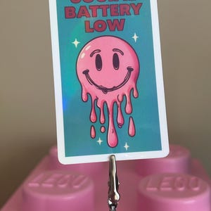 K&ouml;nnte beinhalten: Eine rechteckige Karte mit dem Text "SOCIAL BATTERY LOW" &uuml;ber einem rosa, schmelzenden Smiley-Gesicht. Die Karte wird von einer silbernen Klammer gehalten, und der Hintergrund ist ein rosa Lego-Stein.