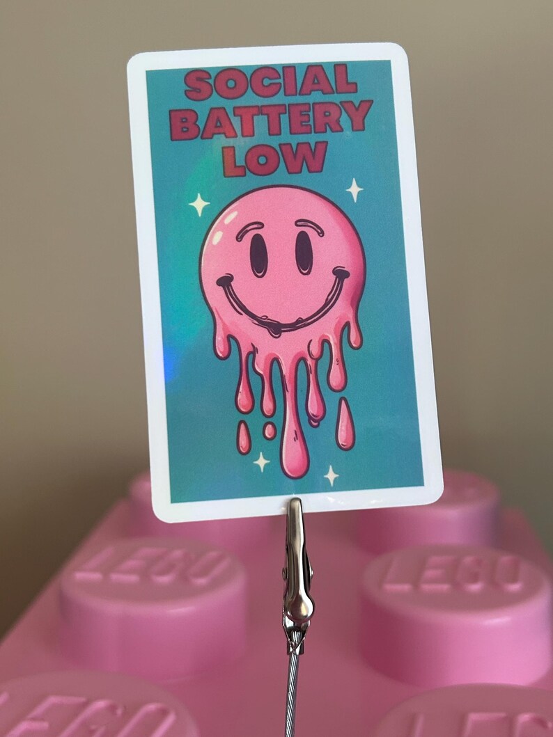 K&ouml;nnte beinhalten: Eine rechteckige Karte mit dem Text "SOCIAL BATTERY LOW" &uuml;ber einem rosa, schmelzenden Smiley-Gesicht. Die Karte wird von einer silbernen Klammer gehalten, und der Hintergrund ist ein rosa Lego-Stein.