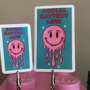 K&ouml;nnte beinhalten: Zwei Karten mit dem Text "SOCIAL BATTERY LOW" und einem rosa schmelzenden Smiley-Gesicht auf t&uuml;rkisblauem Hintergrund. Die Karten werden von silbernen Clips gehalten, die an einem rosa Lego-Stein befestigt sind.