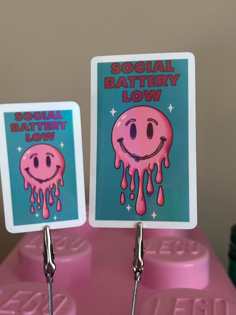K&ouml;nnte beinhalten: Zwei Karten mit dem Text "SOCIAL BATTERY LOW" und einem rosa schmelzenden Smiley-Gesicht auf t&uuml;rkisblauem Hintergrund. Die Karten werden von silbernen Clips gehalten, die an einem rosa Lego-Stein befestigt sind.