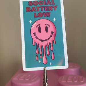 K&ouml;nnte beinhalten: Rechteckiger Aufkleber mit dem Text "SOCIAL BATTERY LOW" in roten Buchstaben. Der Aufkleber zeigt ein rosa, schmelzendes Smiley-Gesicht auf t&uuml;rkisblauem Hintergrund mit wei&szlig;en Sternakzenten. Der Aufkleber wird von einer silbernen Klammer gehalten.