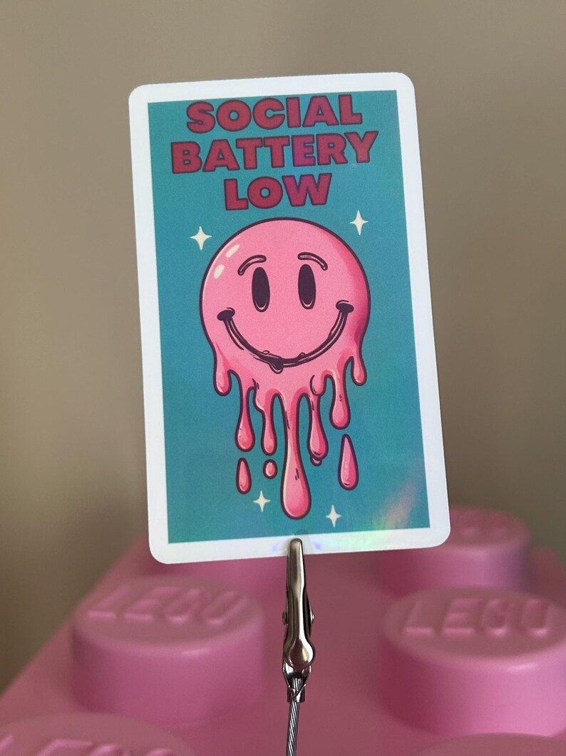 K&ouml;nnte beinhalten: Rechteckiger Aufkleber mit dem Text "SOCIAL BATTERY LOW" in roten Buchstaben. Der Aufkleber zeigt ein rosa, schmelzendes Smiley-Gesicht auf t&uuml;rkisblauem Hintergrund mit wei&szlig;en Sternakzenten. Der Aufkleber wird von einer silbernen Klammer gehalten.