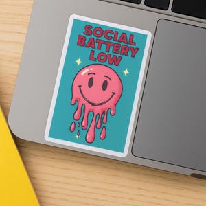 K&ouml;nnte beinhalten: Ein rechteckiger Aufkleber mit dem Text "SOCIAL BATTERY LOW" in roten Buchstaben. Der Aufkleber zeigt ein rosa, schmelzendes Smiley-Gesicht auf t&uuml;rkisem Hintergrund, mit gelben Sternakzenten.
