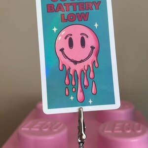 K&ouml;nnte beinhalten: Eine rechteckige Karte mit dem Text "SOCIAL BATTERY LOW" und einem rosa, schmelzenden Smiley-Gesicht. Die Karte wird von einer silbernen Klammer und einem Draht gehalten, vor einem rosa Hintergrund.