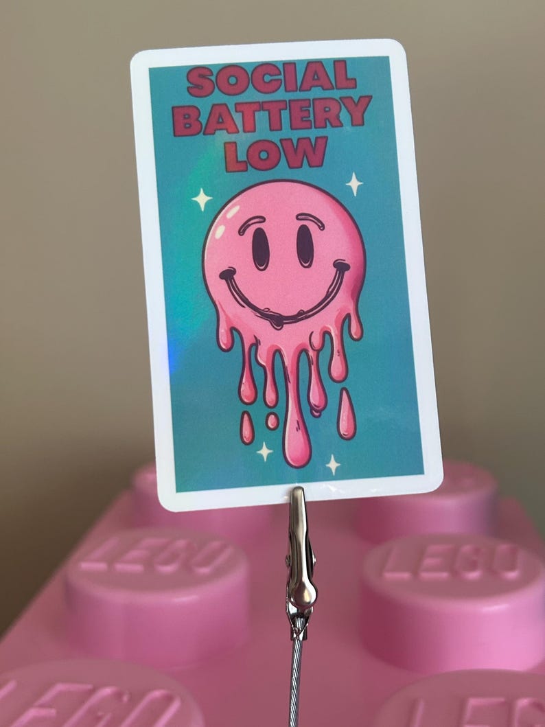 K&ouml;nnte beinhalten: Eine rechteckige Karte mit dem Text "SOCIAL BATTERY LOW" und einem rosa, schmelzenden Smiley-Gesicht. Die Karte wird von einer silbernen Klammer und einem Draht gehalten, vor einem rosa Hintergrund.
