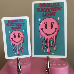 K&ouml;nnte beinhalten: Zwei Karten mit t&uuml;rkisfarbenem Hintergrund und dem Text "SOCIAL BATTERY LOW" in Rot. Jede Karte zeigt ein rosa, schmelzendes Smiley-Gesicht. Die Karten werden von silbernen Clips auf einer rosa Oberfl&auml;che gehalten.
