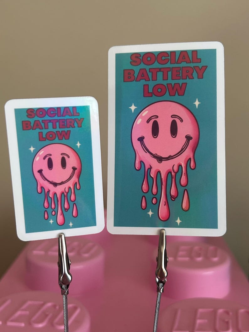 K&ouml;nnte beinhalten: Zwei Karten mit t&uuml;rkisfarbenem Hintergrund und dem Text "SOCIAL BATTERY LOW" in Rot. Jede Karte zeigt ein rosa, schmelzendes Smiley-Gesicht. Die Karten werden von silbernen Clips auf einer rosa Oberfl&auml;che gehalten.