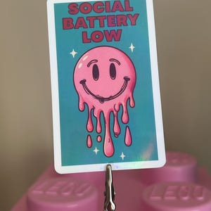 K&ouml;nnte beinhalten: Rechteckiger Aufkleber mit dem Text "SOCIAL BATTERY LOW" in roten Buchstaben. Der Aufkleber zeigt ein rosa, schmelzendes Smiley-Gesicht auf t&uuml;rkisblauem Hintergrund mit wei&szlig;en Sternakzenten. Der Aufkleber wird von einer Metallklammer gehalten.