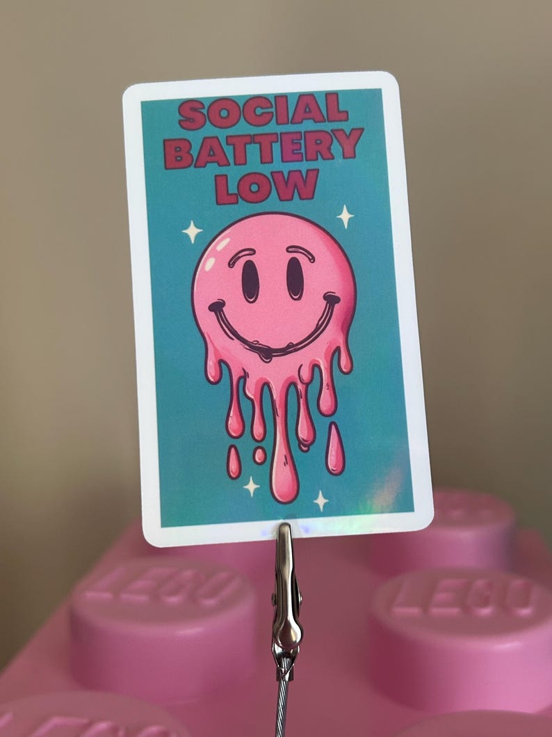 K&ouml;nnte beinhalten: Rechteckiger Aufkleber mit dem Text "SOCIAL BATTERY LOW" in roten Buchstaben. Der Aufkleber zeigt ein rosa, schmelzendes Smiley-Gesicht auf t&uuml;rkisblauem Hintergrund mit wei&szlig;en Sternakzenten. Der Aufkleber wird von einer Metallklammer gehalten.