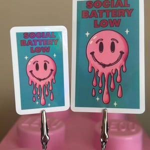 K&ouml;nnte beinhalten: Zwei Karten mit einem rosa schmelzenden Smiley-Gesicht-Design und dem Text "SOCIAL BATTERY LOW" auf t&uuml;rkisfarbenem Hintergrund. Die Karten werden von silbernen Clips gehalten und auf einer rosa Oberfl&auml;che pr&auml;sentiert. Das Design enth&auml;lt wei&szlig;e Sternakzente.