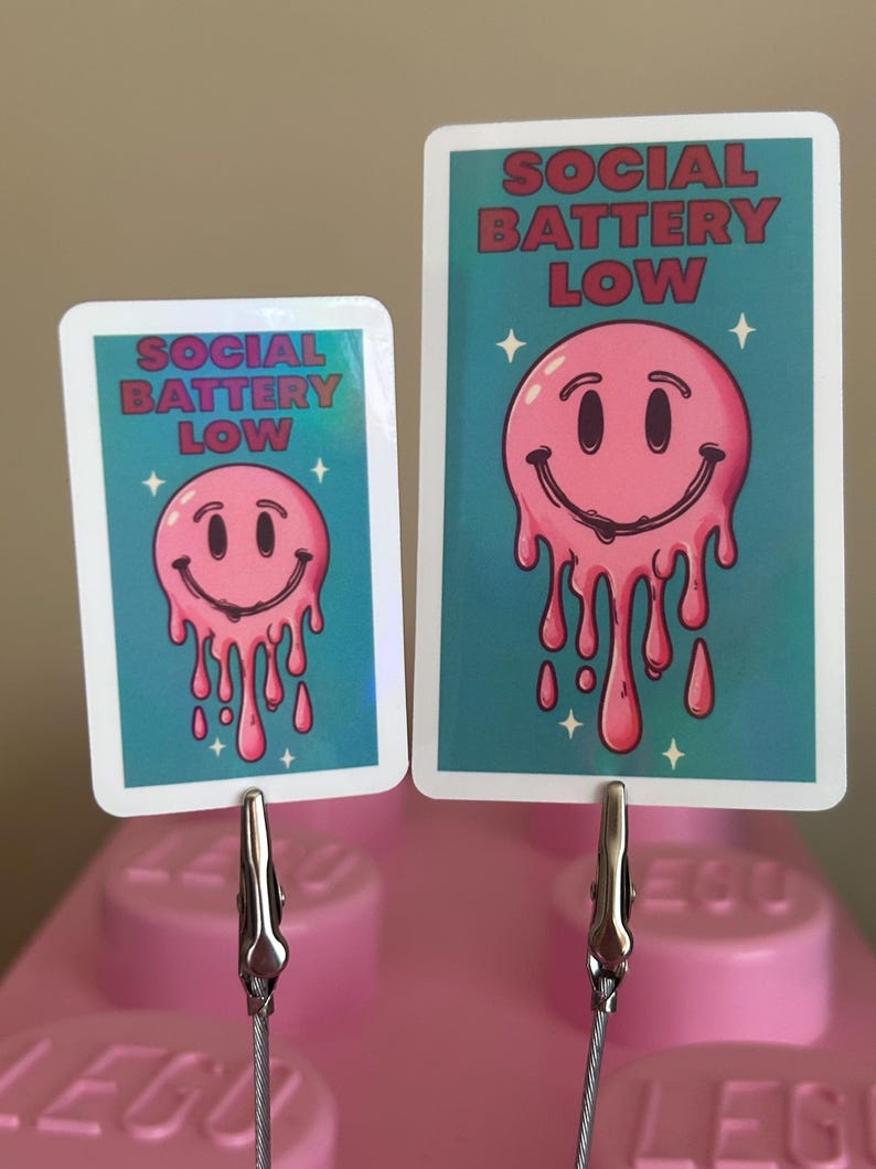 K&ouml;nnte beinhalten: Zwei Karten mit einem rosa schmelzenden Smiley-Gesicht-Design und dem Text "SOCIAL BATTERY LOW" auf t&uuml;rkisfarbenem Hintergrund. Die Karten werden von silbernen Clips gehalten und auf einer rosa Oberfl&auml;che pr&auml;sentiert. Das Design enth&auml;lt wei&szlig;e Sternakzente.