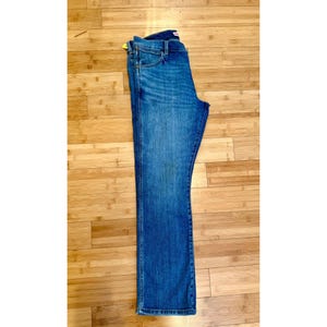Pu&ograve; includere: Jeans di denim blu con un classico design a cinque tasche. I jeans hanno un taglio dritto e un lavaggio medio con sbiadimento sottile. I jeans sono piegati e appoggiati su una superficie di legno.