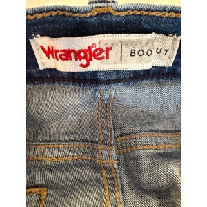 Pu&ograve; includere: Primo piano del retro di jeans blu. L'etichetta del marchio Wrangler &egrave; visibile, con la parola "Wrangler" in rosso e "BOOT" in nero. I jeans hanno cuciture marroni.