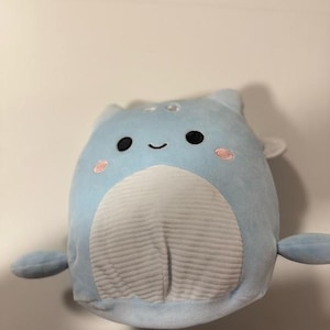 Puede incluir: Un peluche azul claro con forma de ballena. Tiene ojos negros, una boca sonriente y mejillas rosadas. El vientre es blanco con una textura acanalada vertical y tiene pequeñas aletas y cola.