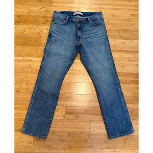 Pu&ograve; includere: Jeans blu in denim con un classico design a cinque tasche. I jeans hanno un taglio dritto e una chiusura con bottone e cerniera. L'etichetta del marchio Wrangler &egrave; visibile in vita. I jeans sono stesi su una superficie di legno.