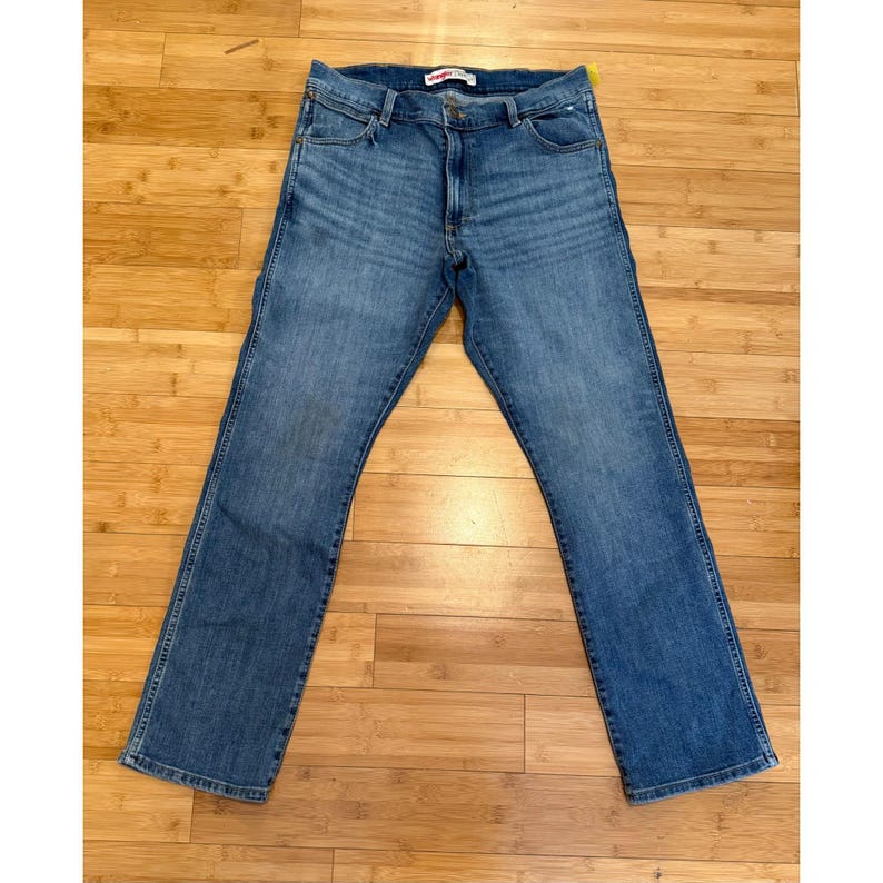 Pu&ograve; includere: Jeans blu in denim con un classico design a cinque tasche. I jeans hanno un taglio dritto e una chiusura con bottone e cerniera. L'etichetta del marchio Wrangler &egrave; visibile in vita. I jeans sono stesi su una superficie di legno.