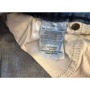 Pu&ograve; includere: Primo piano dell'interno di jeans blu, che mostra l'etichetta di cura. L'etichetta include i numeri RN e CA, la composizione del tessuto e la taglia 34x32. I jeans sono fabbricati in Bangladesh.