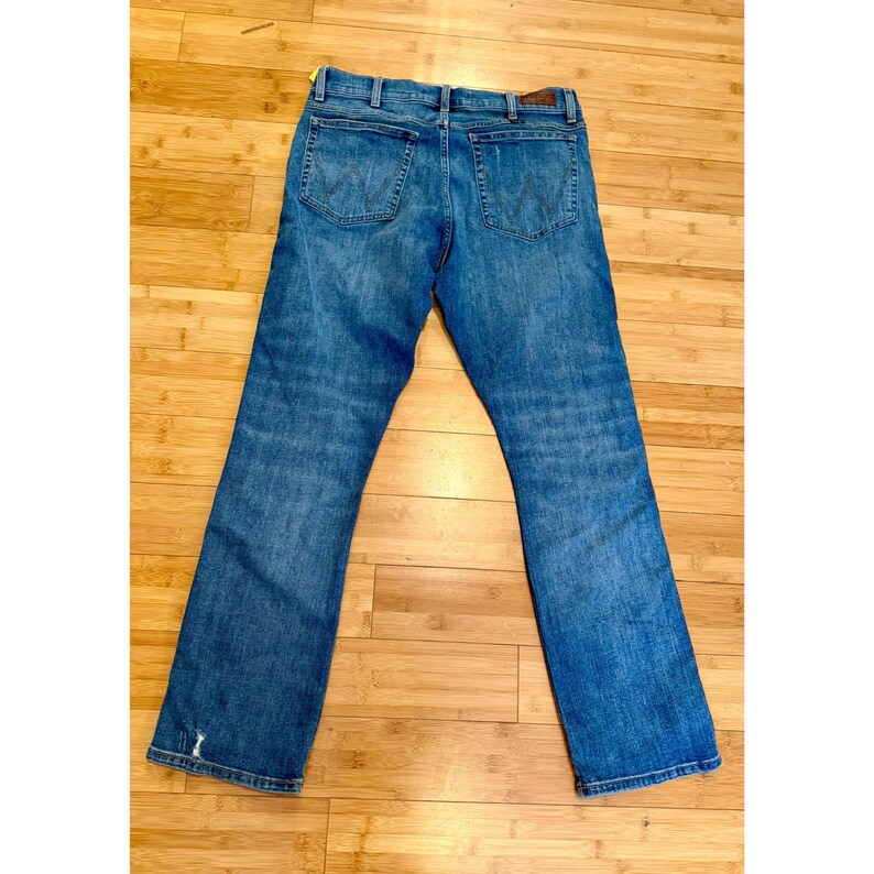 Pu&ograve; includere: Jeans blu in denim con un classico design a cinque tasche. I jeans hanno un taglio dritto e un lavaggio sbiadito. Le tasche posteriori presentano un design a 'W' cucito. I jeans sono stesi su una superficie di legno.
