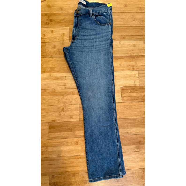 Pu&ograve; includere: Jeans in denim blu con un classico design a cinque tasche. I jeans hanno un taglio dritto e un lavaggio medio. Il capo &egrave; piegato e appoggiato su una superficie di legno. Un'etichetta gialla &egrave; attaccata alla cintura.