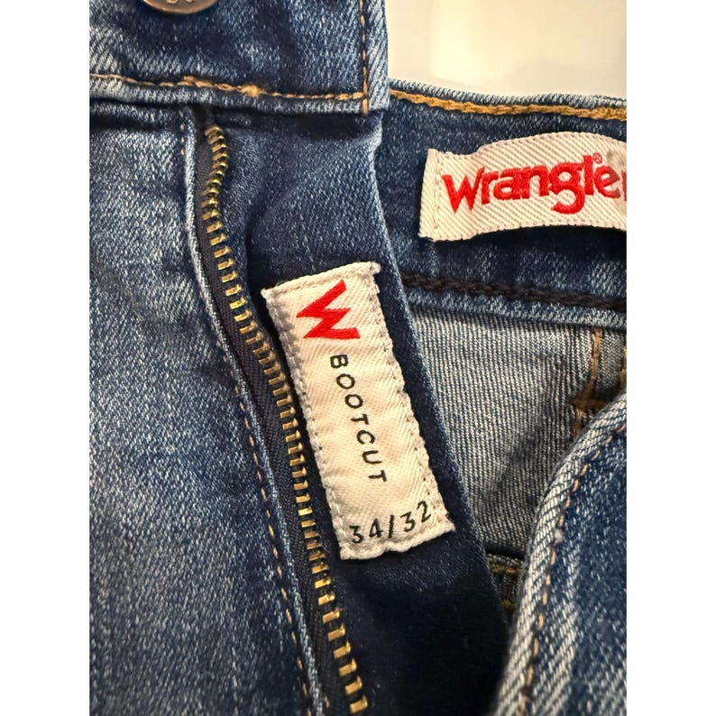 Pu&ograve; includere: Primo piano di jeans bootcut Wrangler in denim blu. I jeans hanno una cerniera e un'etichetta bianca con la scritta "BOOTCUT" e "34/32". Il logo Wrangler &egrave; visibile su un'etichetta separata.