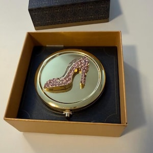 May include: A gold-toned compact mirror with a high-heeled shoe design encrusted with pink rhinestones. The mirror is presented in a gold box with a navy blue interior and a matching lid.