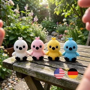 May include: Four small, round, crocheted bird figures in white, light pink, yellow, and light blue. Each figure features a small tuft on top, black eyes, a black beak, and black feet. The figures are arranged in a row on a wooden surface.