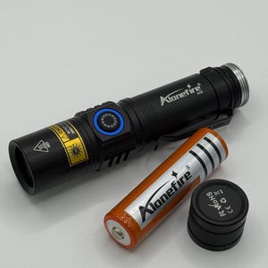May include: Black Alonefire flashlight with a blue power button, a silver clip, and the Alonefire logo. An orange battery and a black cap are next to the flashlight. The flashlight has a warning label.