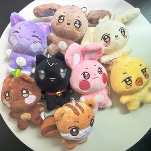 May include: A collection of plush animal keychains in various colors. The keychains include a purple cat, brown dog, black cat, pink bunny, yellow chick, and orange tiger. Each plush has embroidered details and a metal keychain.