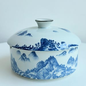 May include: A round ceramic lidded container featuring a white base with a blue hand-painted landscape design. The pattern depicts mountains, trees, and rolling hills wrapping around the body and the lid, which is topped with a small knob.
