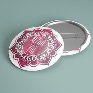 May include: Two white round badges with a decorative floral design in shades of red and pink. The text "ALPHA PHI" is centered on one badge. The other badge shows the pin back. The badges are on a teal background.