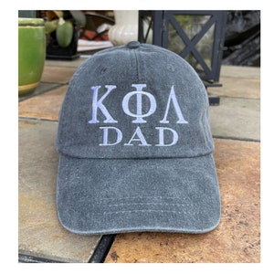 May include: A black, distressed, baseball cap with white embroidered text that reads "KΦΛ DAD".
