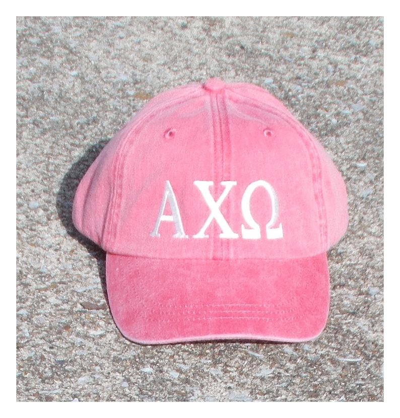 May include: A pink baseball cap with a curved brim. The cap features the Greek letters A, X, and &Omega; embroidered in white on the front. The hat is made of a soft, textured material.