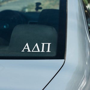 May include: A white vinyl sticker with the letters "ΑΔΠ" on a silver car.