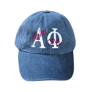 May include: A blue denim baseball cap with white embroidered letters that say "Alpha Phi" in a cursive font. The letters "Alpha" are in red.