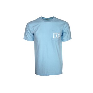 May include: A light blue t-shirt with a pocket on the front. The pocket has the letters "ΠΚΦ" embroidered on it in white.