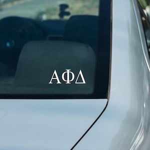 May include: A white vinyl decal with the Greek letters Alpha Phi Delta on a silver car window.