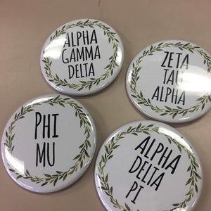 Op de afbeelding: Vier witte ronde knoppen met groene bladerenkransen rond de randen. Elke knop heeft zwarte tekst in het midden. De knoppen lezen "Alpha Gamma Delta", "Zeta Tau Alpha", "Phi Mu" en "Alpha Delta Pi".