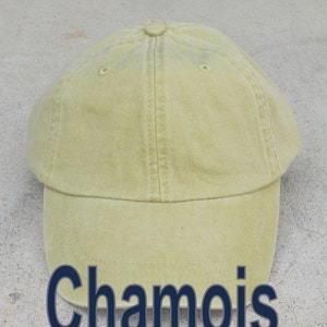 May include: A light yellow baseball cap with the word "Chamois" in dark blue block letters across the bottom. The cap has a curved brim and a button on top. The cap is made of a soft, textured fabric.