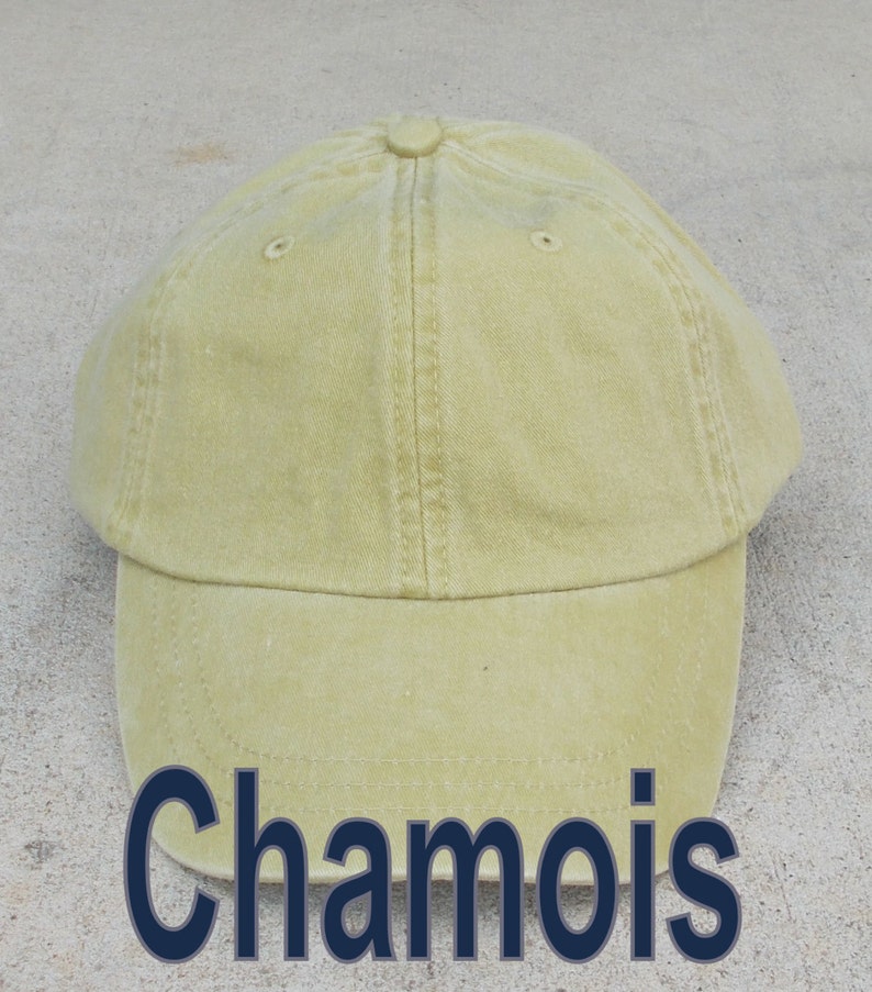 May include: A light yellow baseball cap with the word "Chamois" in dark blue block letters across the bottom. The cap has a curved brim and a button on top. The cap is made of a soft, textured fabric.