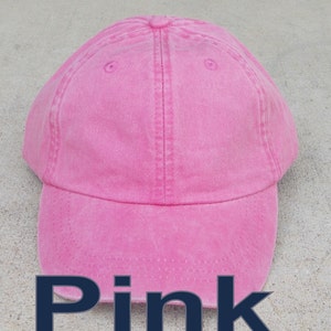 May include: A pink baseball cap with a curved brim. The cap is a solid, vibrant pink color and features a classic six-panel design with a top button and adjustable strap. The word "Pink" is displayed in a dark blue font at the bottom.