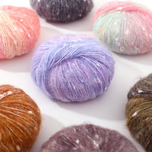 May include: Close-up of several colorful yarn balls. The yarn balls are in shades of pink, purple, brown, and gray, with white flecks throughout. The yarn appears soft and fluffy, ideal for knitting or crocheting projects.