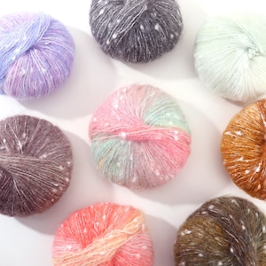 May include: A collection of colorful yarn balls in various shades, including purple, pink, and brown, with white speckles. The yarn appears soft and fluffy, ideal for knitting or crocheting projects. The balls are arranged on a white surface.
